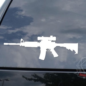 May include: White vinyl decal of a rifle with a scope, affixed to a car window. The image shows the silhouette of the firearm, with details of the barrel, stock, and scope visible. The background is a cloudy sky.