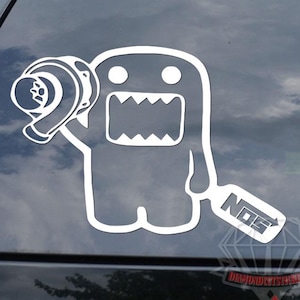 May include: White vinyl car decal featuring a cartoon character holding a turbocharger and a NOS logo. The character has a large mouth and two eyes. The decal is applied to a black car window.
