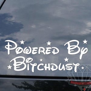 May include: White vinyl decal with the text "Powered By Bitchdust" in a whimsical font, accented with stars. The decal is applied to the rear window of a black vehicle.