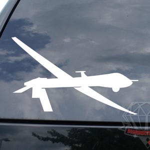 May include: White vinyl decal of a drone aircraft, affixed to a black vehicle window. The drone features a detailed wing and body design. The background shows a cloudy sky.