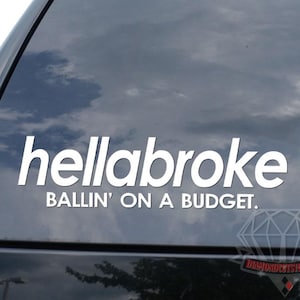 May include: White vinyl decal with the text "hellabroke" above "BALLIN' ON A BUDGET." applied to a car window. The background shows a cloudy sky reflected in the glass.