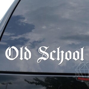 May include: White "Old School" decal on a car window. The text is in a gothic font. A diamond-shaped logo is visible at the bottom right. The background shows a cloudy sky.