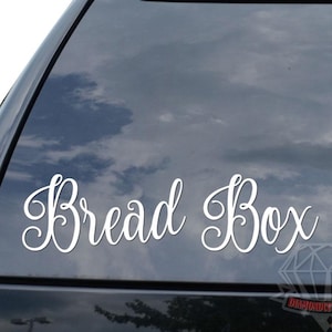 May include: A white cursive "Bread Box" decal is affixed to the rear window of a black vehicle. The decal is in a stylish font. The sky is visible through the window. A diamond-shaped logo is at the bottom.