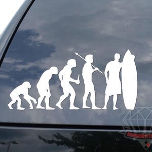 May include: White vinyl decal depicting the evolution of man, culminating in a surfer with a surfboard. The decal is applied to a black vehicle window, showcasing a humorous take on human development and surfing.