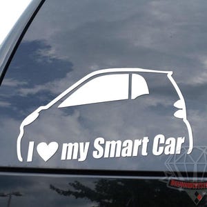 May include: White vinyl car decal featuring a silhouette of a Smart Car with the text "I ♥ my Smart Car". The decal is applied to the rear window of a black vehicle. The sky is visible through the window.