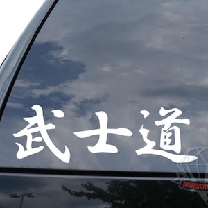 May include: White vinyl decal with Japanese characters on a car window. The characters read "Bushido", the way of the warrior. The decal is applied to the rear window of a black vehicle, with a cloudy sky reflected in the glass.