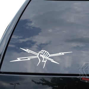 May include: White vinyl decal of a raised fist holding two lightning bolts. The decal is on a car window.