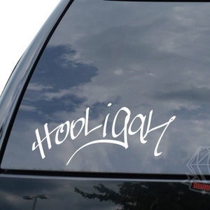 May include: A white vinyl decal with the word "Hooligan" written in a stylized font. The decal is applied to the rear window of a black car.