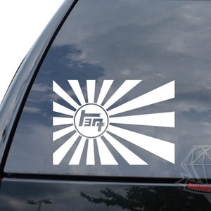 May include: White vinyl decal of a Japanese rising sun with the Toyota logo in the center. The decal is on a black car window.