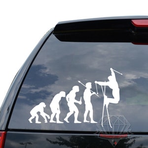 May include: White vinyl car decal depicting the evolution of skiing, from ape to skier. The decal shows a series of figures, each representing a stage in the evolution, culminating in a skier mid-air. The decal is applied to a black car window.