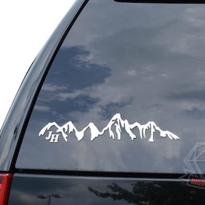 May include: White vinyl decal of a mountain range with the initials "JH" in the foreground. The decal is applied to the rear window of a black SUV.