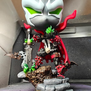 Peut inclure: Figurine de Spawn, principalement noire, rouge et blanche, avec une cape rouge et des pointes, debout sur une base en pierre. Il tient une épée et des flammes vertes. La tête est grise avec des accents blancs et des yeux verts.