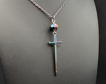 Puede incluir: Un collar de cadena en tono plateado con un colgante en forma de espada suspendido bajo una cuenta única facetada con brillo iridiscente. El colgante de espada tiene un acabado metálico con sutiles tonos arcoíris, una guarda y una punta afilada.