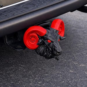 Pode incluir: Cobertura de engate de reboque com cabeça de carneiro preta com chifres vermelhos e olhos vermelhos brilhantes. A cabeça de carneiro detalhada é montada em um para-choque preto. Expressão ameaçadora.