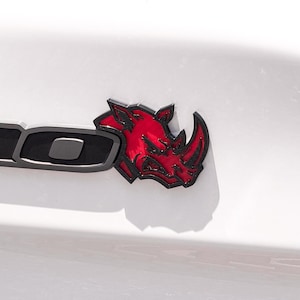 May include: A car emblem featuring a stylized red rhinoceros head with black outlines, positioned next to a partial black letter on a white surface. The design shows a sharp, aggressive rhino profile with detailed facial features and a prominent horn.