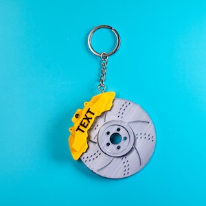 May include: A silver brake rotor keychain with a yellow brake pad attached. The brake pad has the word "TEXT" printed on it.
