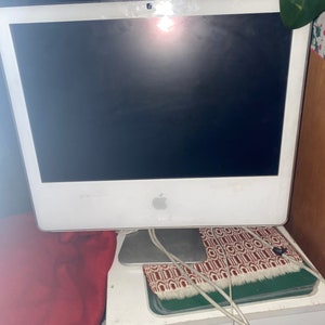 May include: A white iMac computer with a black screen, sitting on a white desk with a red and white patterned mat and a green mat underneath. The computer is connected to a power cord and a USB cable.