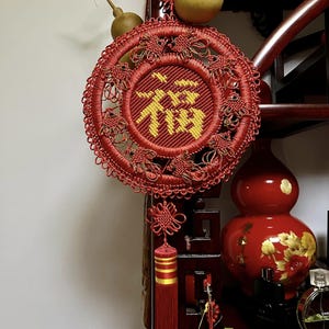 Op de afbeelding: Een rode Chinese knoop met een gouden kwast en het Chinese karakter voor "geluk" geborduurd in gouden draad. De knoop hangt aan een houten plank met pompoenen en andere decoraties.