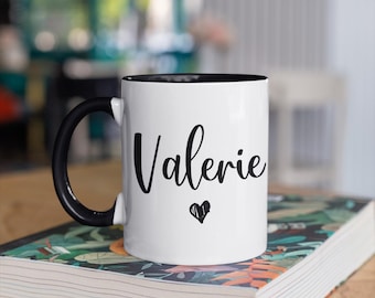 Könnte beinhalten: Weiße Keramiktasse mit schwarzem Henkel und schwarzem Rand. Die Tasse trägt den Namen "Valerie" in schwarzer Kursivschrift und ein kleines schwarzes Herz unter dem Namen.