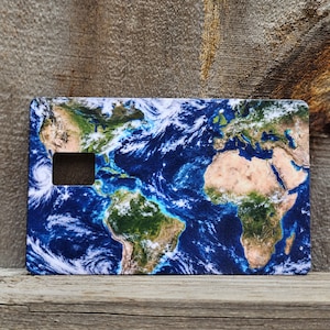 May include: A rectangular credit card skin featuring a high-resolution satellite view of the Earth. The design displays vibrant blue oceans, green and tan landmasses, and swirling white cloud formations. A rectangular cutout is positioned on the left.