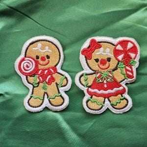 May include: Two embroidered gingerbread man patches. One holds a red and white swirled lollipop and wears a red scarf. The other wears a red dress and bow, and holds a red and white swirled lollipop and candy cane. Both have tan bodies, white frosting, and black eyes.