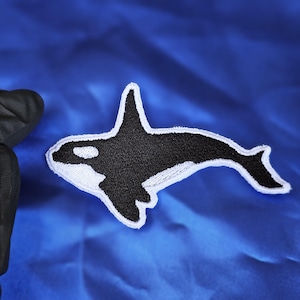 May include: Embroidered patch featuring a black and white orca whale design. The patch has a white border and is set against a vibrant blue background. Ideal for jackets, bags, or other fabric items.