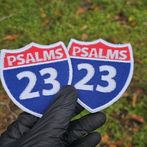May include: Two embroidered patches in the shape of highway signs. Each patch features a red border, the word "PSALMS" in white, and the number "23" in white on a blue background. The patches are held against a blurred green background.