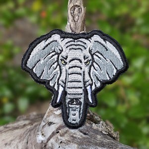 May include: Embroidered elephant patch with gray and white details, outlined in black. The elephant's eyes are yellow. The patch is displayed on a piece of driftwood against a blurred green background.
