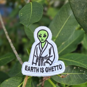 May include: Embroidered patch featuring a green alien wearing a white robe with a black belt. The patch has the text "EARTH IS GHETTO" in black letters on a white background. The patch is surrounded by green leaves.