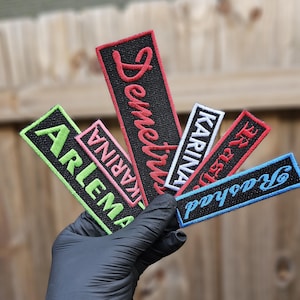 May include: Five embroidered patches with different names on them. The patches are black, red, pink, white, and blue. The patches are held in a black gloved hand.