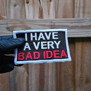 May include: A black patch with white stitching and the text "I HAVE A VERY BAD IDEA" in white and red lettering.