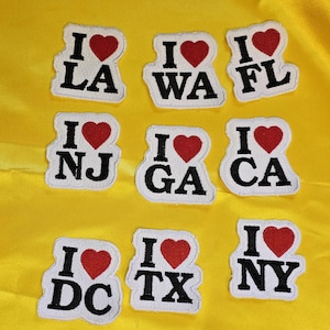 May include: Nine white felt patches with red hearts and black lettering. The patches read "I love LA", "I love WA", "I love FL", "I love NJ", "I love GA", "I love CA", "I love DC", "I love TX", and "I love NY".