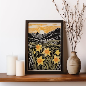 Op de afbeelding: Een ingelijste kunstdruk met een zwart-geel landschap met narcissen. Het kunstwerk toont een zonsopgang boven glooiende heuvels en bergen. De print wordt tentoongesteld op een houten plank met kaarsen en een vaas.
