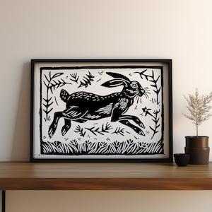 May include: Black and white framed art print featuring a running hare. The linocut style print depicts the hare in motion amidst stylized foliage. The artwork is displayed on a wooden surface, with a small plant and bowl nearby.