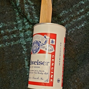 May include: A Budweiser beer can with a wooden handle attached. The can is white with red and blue accents and the word "GENUINE" in red. The handle is made of light-colored wood.