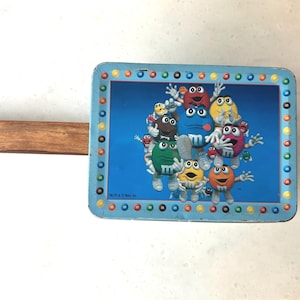 May include: Vintage blue metal lunchbox featuring a colorful graphic of M&M's candies. The lunchbox has a rectangular shape with a wooden handle attached. The image shows the lunchbox on a white surface.