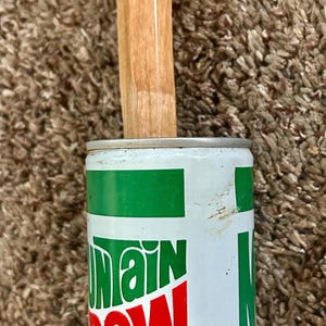 May include: A Mountain Dew soda can with a 355 ml capacity label, featuring white and green branding with red text. A wooden ruler is positioned vertically against the top of the can, showing measurement markings including the numbers 10 and 11.