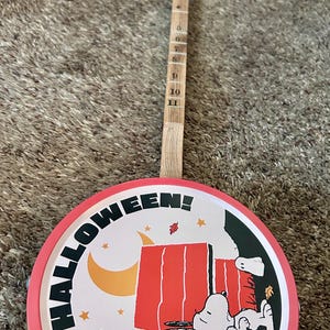 May include: A round, red and white banjo with a wooden neck. The banjo head features a Snoopy Halloween design with the words "HAPPY HALLOWEEN!" in a circular pattern around the image.