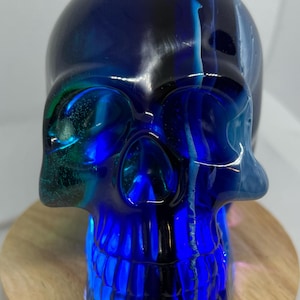 Può includere: Un teschio di vetro blu con striature bianche e nere, posto su una base di legno. Il teschio è illuminato dall'interno, proiettando un bagliore blu.