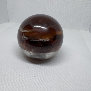 Puede incluir: Una esfera decorativa de vidrio con un interior marrón y ámbar, que descansa sobre una base plateada. La esfera tiene una apariencia veteada con remolinos blancos y ámbar. La base es de color plateado metálico.