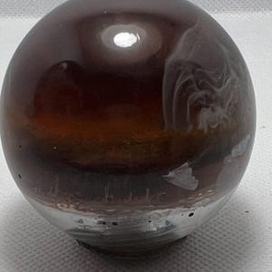 May include: A brown and white glass sphere with swirling patterns inside. The sphere is smooth and shiny.