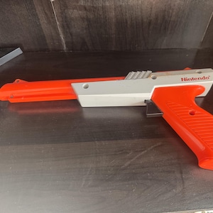 May include: A red and white Nintendo Zapper light gun with a black trigger and a cord. The gun is on a dark wood surface.