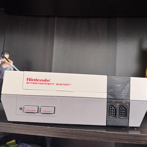 May include: A white Nintendo Entertainment System console with a black controller port and two buttons labelled "Power" and "Reset".