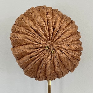 May include: A round, brown, woven hat with a decorative design. The hat is on a metal stand.