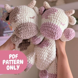 May include: Two crocheted cow plushies, one white with pink spots and one brown with white spots. The plushies are held by hands and are facing the camera. The plushies are made with a soft, fluffy yarn. The text "PDF PATTERN ONLY" is visible in the lower left corner of the image.