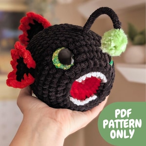 Puede incluir: Un pez rape negro tejido a ganchillo con aletas rojas y negras, ojos grandes y un señuelo de pompón verde. El pez tiene una boca muy abierta con dientes blancos y un interior rojo. La imagen incluye el texto "PDF PATTERN ONLY".