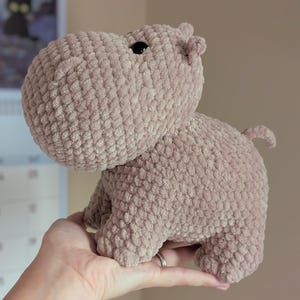 May include: A soft, brown crocheted hippopotamus plush toy. The hippo is standing on its four legs and has a small, black eye.