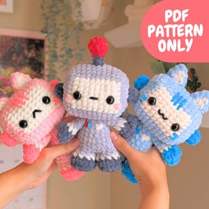 Könnte beinhalten: Drei handgefertigte Häkeltier-Plüschtiere in Rosa, Lavendel und Blau. Das rosa Spielzeug ist eine Katze, das Lavendelspielzeug ist ein Roboter und das blaue Spielzeug ist eine Katze. Jedes Spielzeug hat schwarze Augen und ein niedliches Design. Das Bild enthält den Text "PDF PATTERN ONLY".