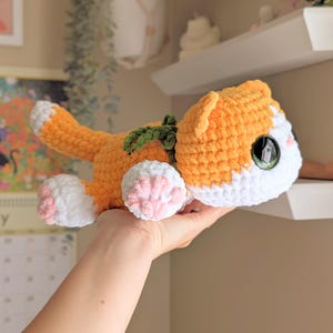 May include: A crocheted orange and white cat plush toy with green detailing. The handmade cat features pink paw pads and large, detailed eyes. The cat is lying down, with its tail curled up. A unique, handcrafted item.