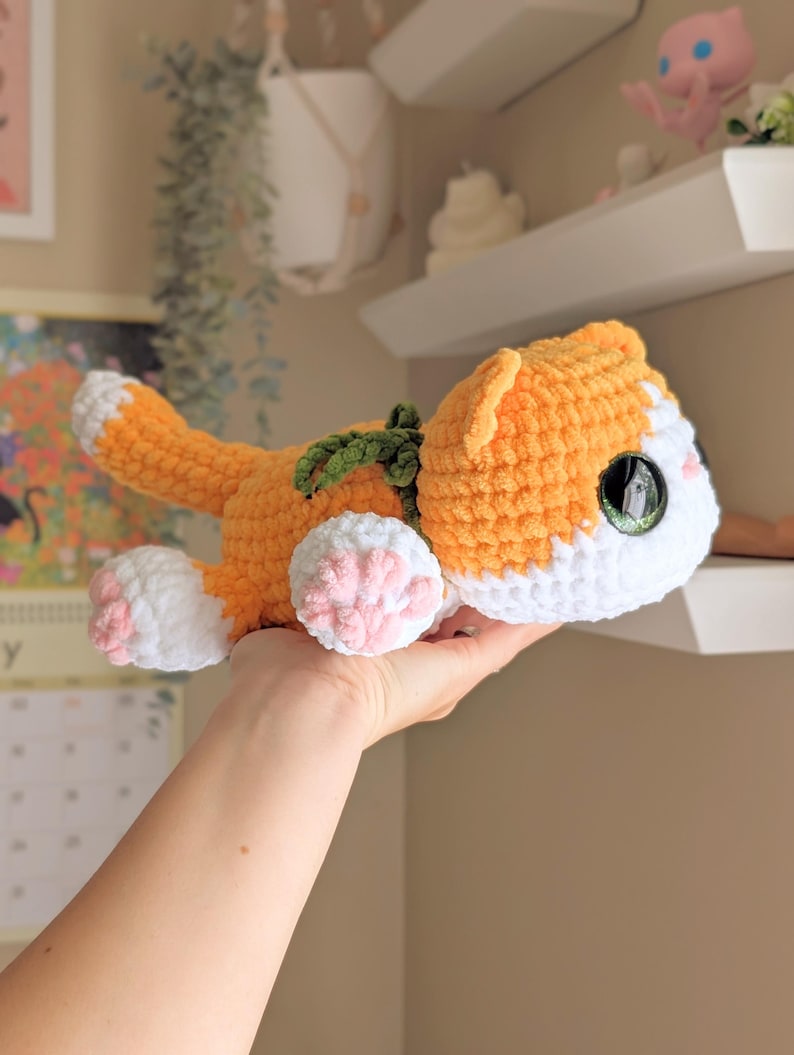 May include: A crocheted orange and white cat plush toy with green detailing. The handmade cat features pink paw pads and large, detailed eyes. The cat is lying down, with its tail curled up. A unique, handcrafted item.